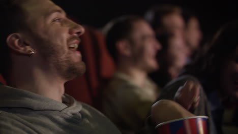 Young-guy-with-earring-in-his-ear-eating-popcorn-laughing-when-watching-movie