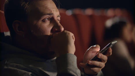 Yawning-man-using-smartphone-in-cinema.-Young-man-watching-boring-film