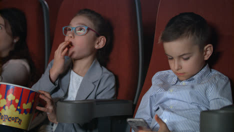 Little-girl-eating-popcorn-in-movie-theater.-Boy-using-smartphone-at-cinema