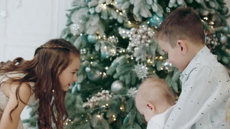 Three-cute-kids-walking-around-christmas-tree-in-living-room.