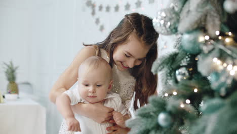 Closeup-happy-girl-holding-kid-near-christmas-tree-in-modern-house.