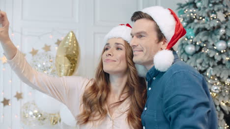 Smiling-couple-making-selfie-in-santa-hats-near-christmas-tree.