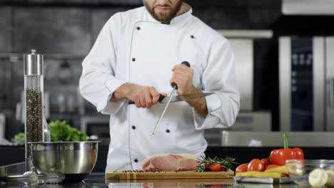 Chef-preparing-to-cook-meat-at-kitchen.-Closeup-chef-hands-sharpening-knife.