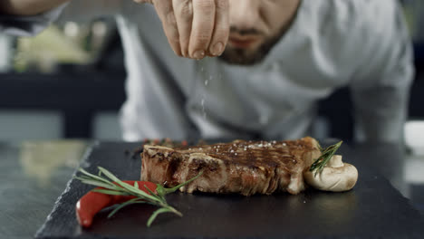 Chef-salt-frying-meat-at-griddle.-Closeup-man-hands-salt-steak-in-slow-motion.