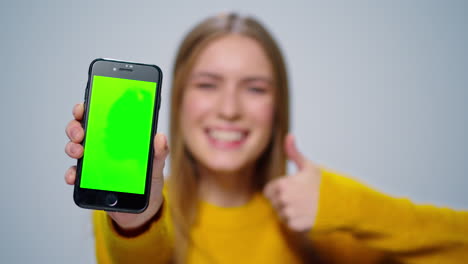 Smiling-woman-showing-smartphone-with-green-screen-in-studio.-Girl-thumbs-up