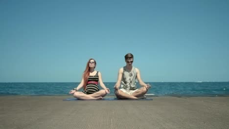 Young-couple-meditating-sitting-on-pier-by-beach.-Holidays-with-yoga-classes