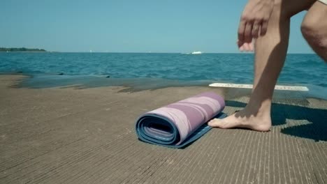 Guy-spreading-yoga-mat-by-sea.-Young-man-preparing-for-meditation-by-sea