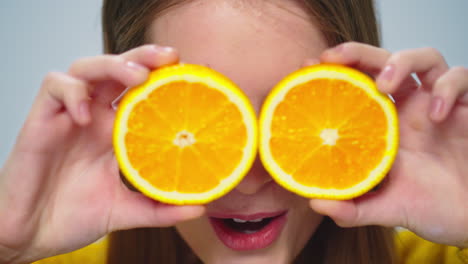 Closeup-cheerful-woman-having-fun-with-two-orange-slices-at-camera-in-studio.