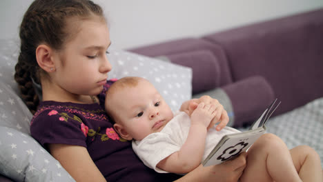 Pretty-girl-reading-kid-book-with-baby-at-home.-Sister-holding-baby-brother