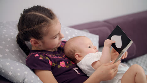 Little-brother-reading-book-with-sister.-Newborn-baby-in-sister-embrace