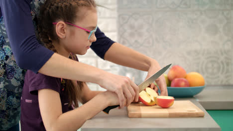 Mother-help-daughter-to-cut-apple-on-cutting-board.-Mom-hand-cooking-with-kid