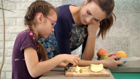 Girl-eating-apple-with-mother-on-kitchen.-Girl-cutting-apple-with-knife