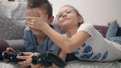 Siblings-enjoy-video-game-at-home.-Girl-close-eyes-boy.-Kids-have-fun-together