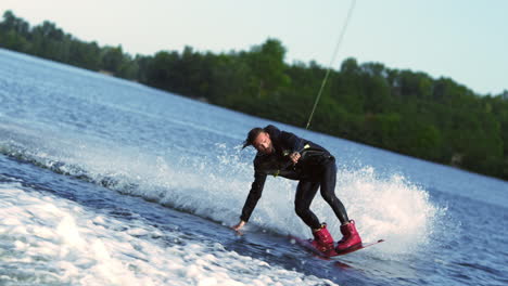 Guy-rushing-along-forest-lake-on-wakeboard-touching-water-surface-by-hand